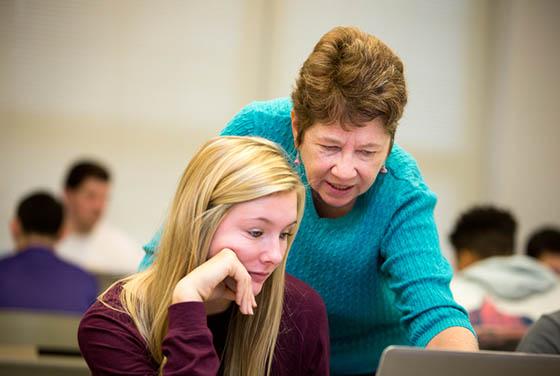 Photo of an older female faculty member leaning over a Chatham University student and pointing to a computer